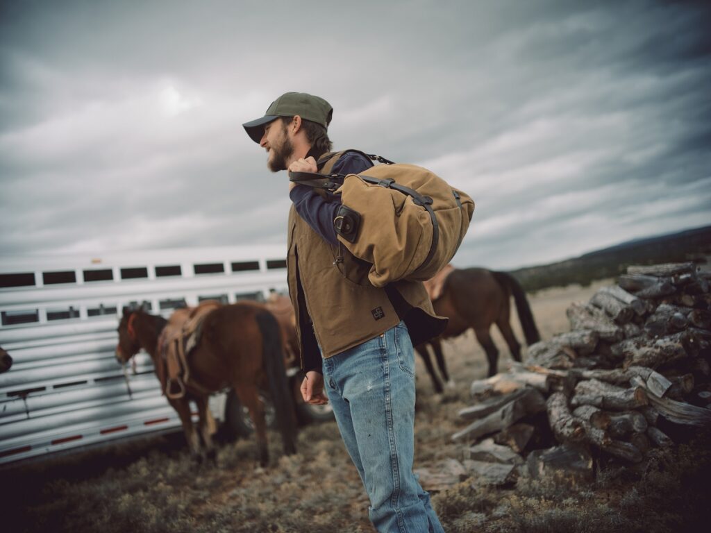 Man holding a Filson duffle bag with horses in the background.